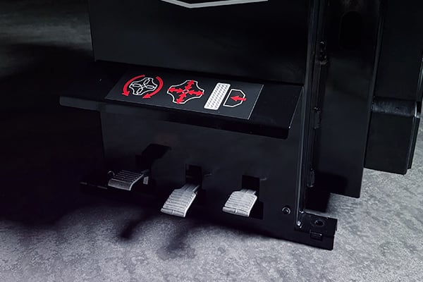 Tres pedales metálicos (acelerador, freno, embrague) montados en una carcasa negra de simulador de conducción, con pictogramas rojos y blancos encima y suelo alfombrado gris.