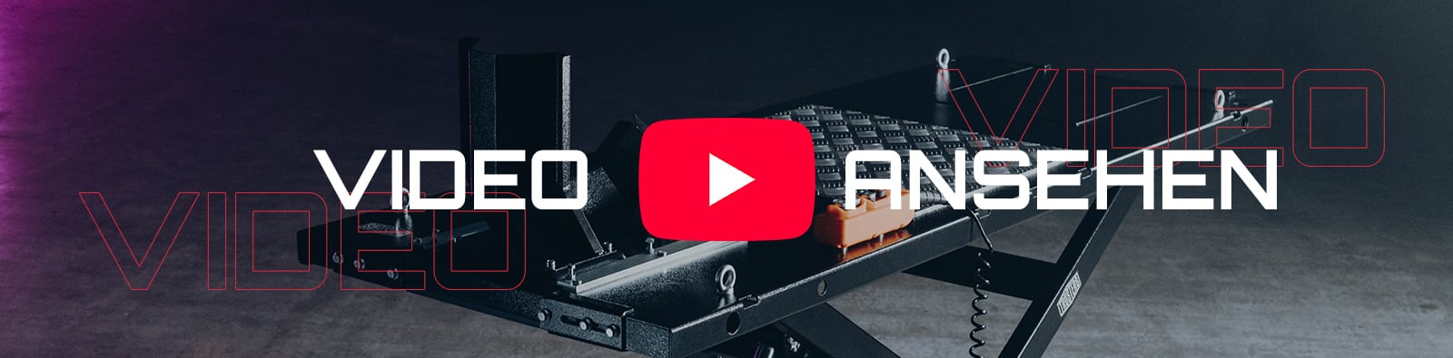 Schräg abgebildete Hebebühne aus schwarzem Metall auf dunklem Betonboden, orange Steuerbox mit Spiralkabel, großer roter Play-Button in der Bildmitte, weiße Schrift VIDEO ANSEHEN und rote konturierte Schrift VIDEO im Hintergrund