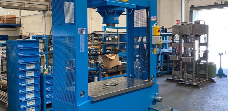 Blaue Werkstattpresse mit großem Metallarbeitstisch und rundem Ausschnitt, im Hintergrund Regale, Schubladenschrank mit blauen Boxen, weitere Maschinen und geöffnete Tür mit Tageslicht