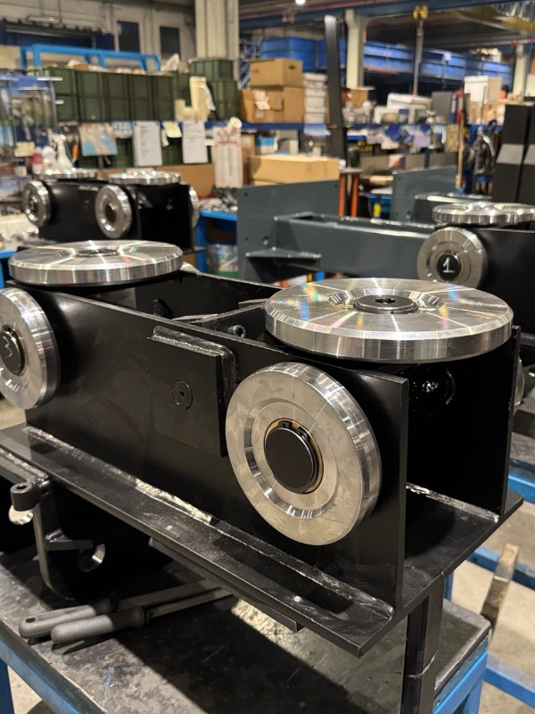 Schwarz lackiertes Metallgestell mit mehreren glänzenden silbernen Laufrädern auf Werkbank, umgeben von Regalen, Kartons und Maschinen in einer Fabrikhalle