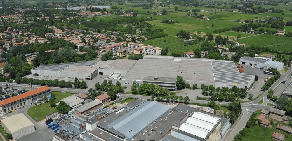Luftaufnahme eines großen Industrie- und Lagerkomplexes mit mehreren Flachdachhallen, Zufahrtsstraßen und Parkplätzen, umgeben von Bäumen, Wohnhäusern und weiten grünen Feldern im Hintergrund