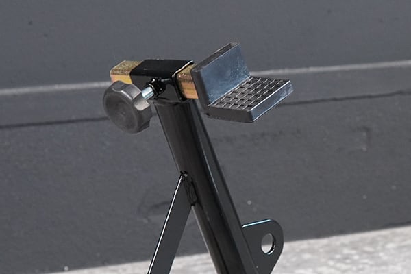 Close-up of a black metal adjustable support bracket with a small textured platform, knurled adjustment knob and triangular brace against a gray wall background