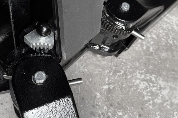 Close-up of two black painted metal hinge brackets with hex bolts, exposed metal pins and a small toothed gear mechanism on a textured concrete floor