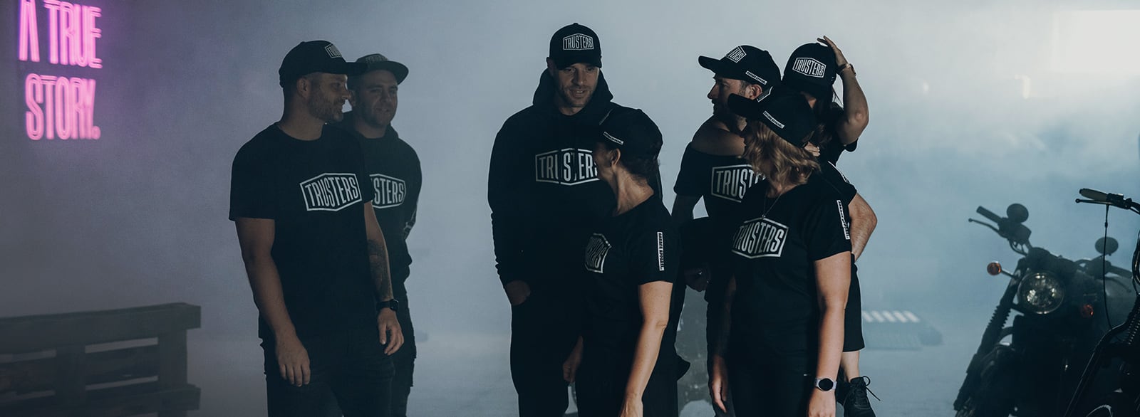 Gruppe von Menschen in schwarzen TRUSTERS-Shirts und Caps steht locker in nebliger Halle neben einem Motorrad und einer Holzpalette; links leuchtet in Neon A TRUE STORY.