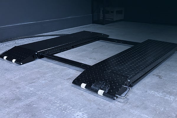 Niedrigprofil-Autohubbühne aus schwarzem Riffelblech: zwei breite Trägerplatten mit weißen Rollen an den vorderen Enden, verbunden durch einen Metallrahmen, auf grauem Betonboden in einer Garage