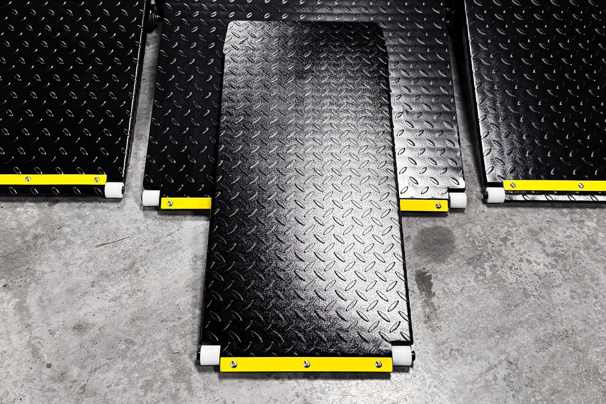 Black diamond-plate metal ramp panels on a concrete floor, arranged in three connected sections with yellow safety strips and white roller end caps