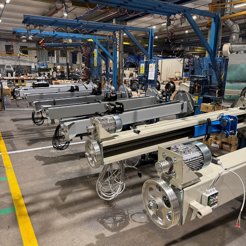 Industrielle Montagehalle mit mehreren parallel aufgebauten Antriebsachsen: lange Metallprofile mit Elektromotoren, großen Riemenscheiben, Kabelbündeln und blauen Montagegestellen, Werkzeugtafeln und Kartons im Hintergrund
