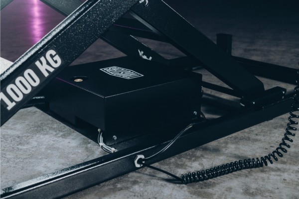 Base metálica de una plataforma elevadora tipo tijera con la inscripción 1000 KG, caja negra de control con cables y cable en espiral sobre suelo de hormigón, iluminación tenue con reflejos morados