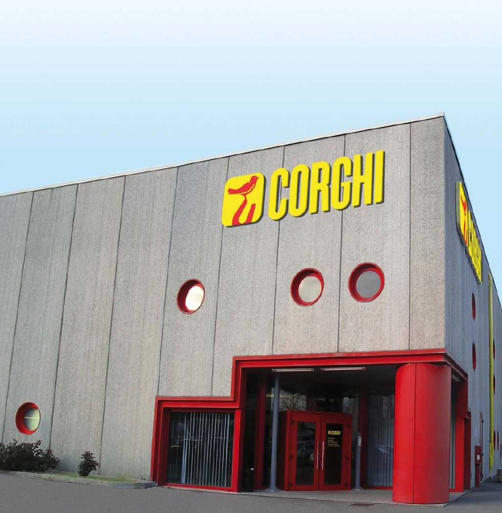 Graue Industriegebäudefassade mit roten runden Bullaugenfenstern, rotem Eingangsportal und gelbem CORGHI-Logo über dem Eingang