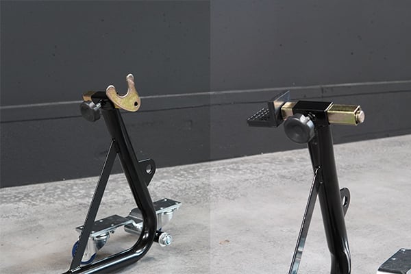 Two black metal motorcycle paddock stands with forked top hooks and small rolling bases, positioned on a concrete floor against a gray wall