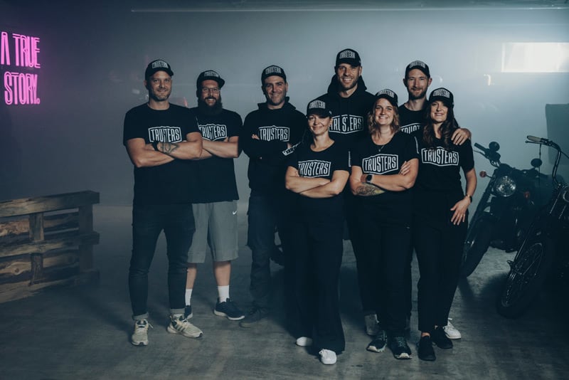 Acht Personen in schwarzer Teamkleidung und Caps mit Schriftzug TRUSTERS, stehen lächelnd mit verschränkten Armen in einer düsteren Garage/Loft-ähnlichen Halle neben Motorrädern und Holzpaletten, neonrosa Leuchtschrift an der Wand