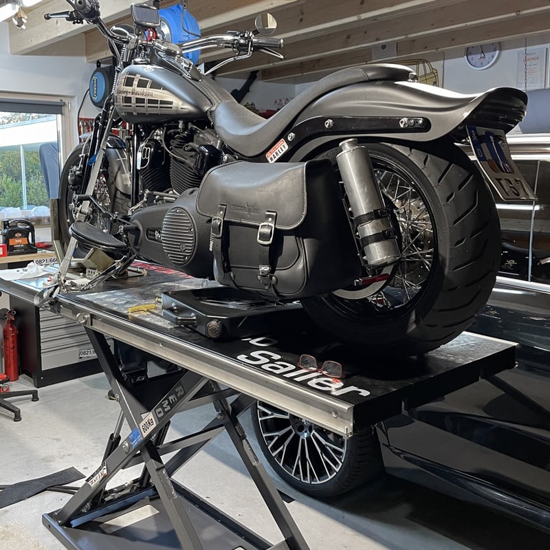 Schwarzes Cruiser-Motorrad mit Ledersatteltasche auf Hebebühne in Werkstatt, sichtbares Hinterrad, Auspuff und Werkzeuge