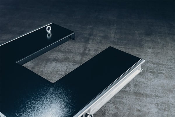 Schwarze, glänzende L-förmige Plattform oder Flightcase-Deckel mit großer rechteckiger Aussparung, Metallkante und Ösenschraube an einer Ecke, auf grauem Betonboden liegend