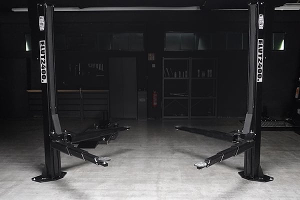 Two-post automotive lift with extended arms centered in a dim industrial garage, concrete floor and shelving and equipment against the back wall.