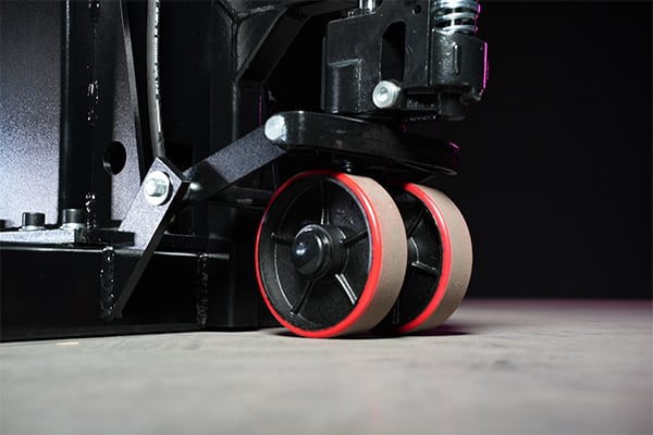 Close-up of dual red-rimmed caster wheels attached to a black industrial pallet jack on a concrete floor