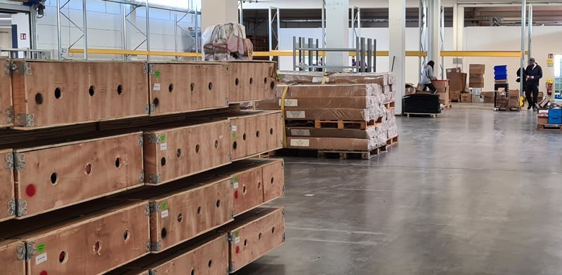 Lagerhalle mit gestapelten Holzkisten links, verpackten Paletten in der Mitte, offenem Lagerraum, Betonboden, hohen Hallenstützen und zwei Personen im Hintergrund