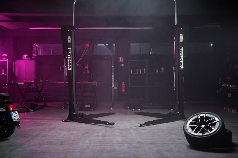 Dunkle Autowerkstatt mit leerer Hebebühne in der Bildmitte, pinkes Licht, Reifen mit Felge rechts im Vordergrund, Roller links, Werkbänke und Regale im Hintergrund, leicht verrauchte Atmosphäre