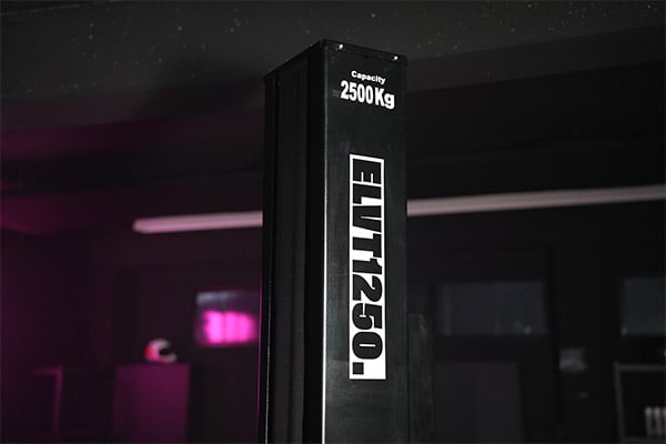 Black vertical lift column labeled ELIT1250. and Capacity 2500Kg in a dim garage with pink ambient light and blurred cabinets and windows