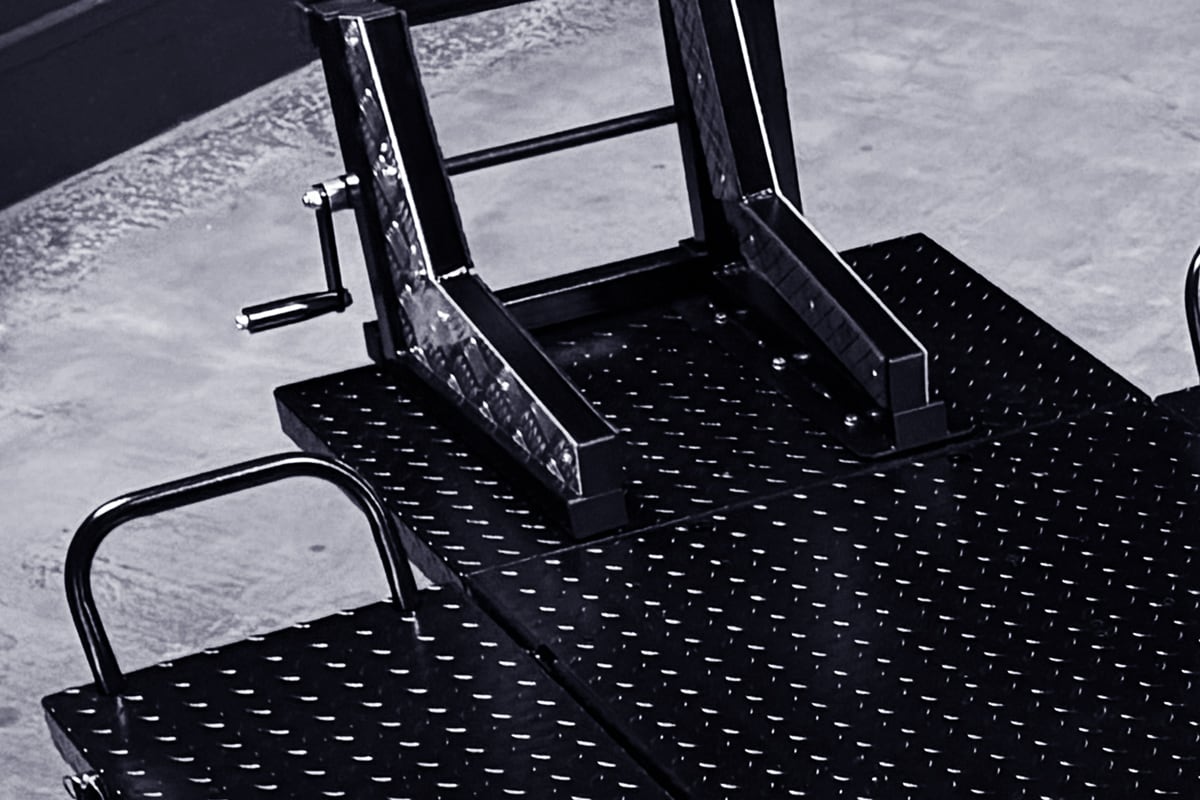 Close-up of black diamond-plate metal platform with welded tubular frame, manual crank handle and curved grab bars on a concrete floor