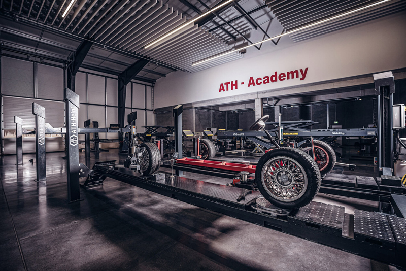 Innenaufnahme einer modernen Kfz‑Schulungswerkstatt mit Fahrzeugrahmen auf Hebebühnen, sichtbaren Rädern und Werkzeugen, poliertem Betonboden und der Aufschrift ATH - Academy an der Wand