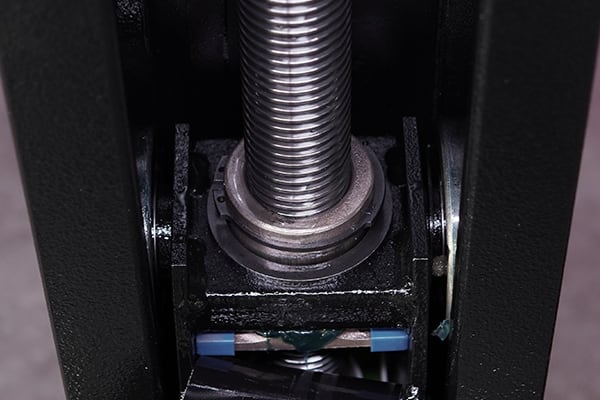 Nahaufnahme einer vertikal eingebauten Gewindespindel mit Metallmutter in schwarzem Rahmen, sichtbaren Lagern, Schmierfett und kleinen blauen Kunststoffteilen