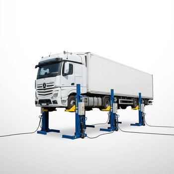 Weißer Lkw mit geschlossenem Kofferaufbau auf vier blauen Hebebühnen angehoben, alle Räder vom Boden abgehoben, Hebevorrichtungen und Anschlusskabel sichtbar vor hellem Hintergrund