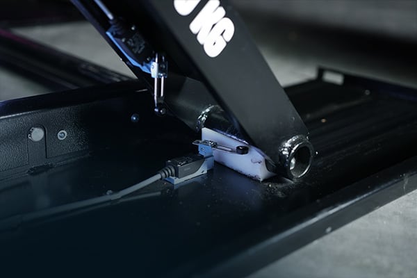 Nahaufnahme eines schwarzen Metallarms mit rundem Gelenk und sichtbarer Schweißnaht, befestigt auf einer flachen schwarzen Basisplatte; ein USB-Kabel ist an einen kleinen Adapter angeschlossen, diffuse blaue Beleuchtung