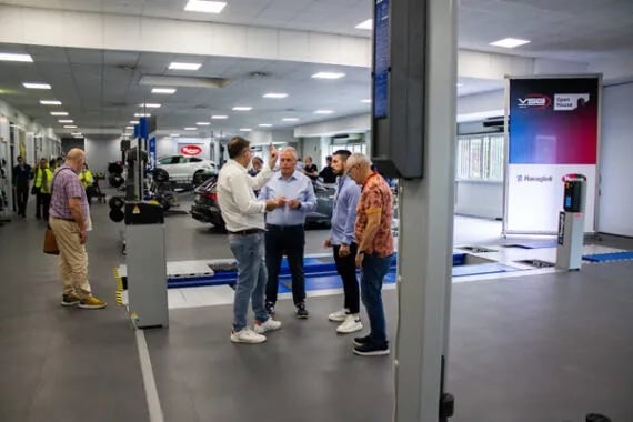 Moderne Autowerkstatt-Servicehalle mit Hebebühnen, geparkten Fahrzeugen und Werkstattausrüstung; fünf Männer in lockerer Kleidung stehen in der Hallenmitte im Gespräch, weitere Mitarbeiter im Hintergrund