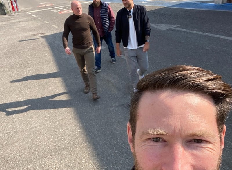 Nahes Selfie eines lächelnden Mannes im Vordergrund, drei Männer gehen hinter ihm auf einem sonnigen Asphaltplatz auf die Kamera zu, lange Schatten und legere Kleidung