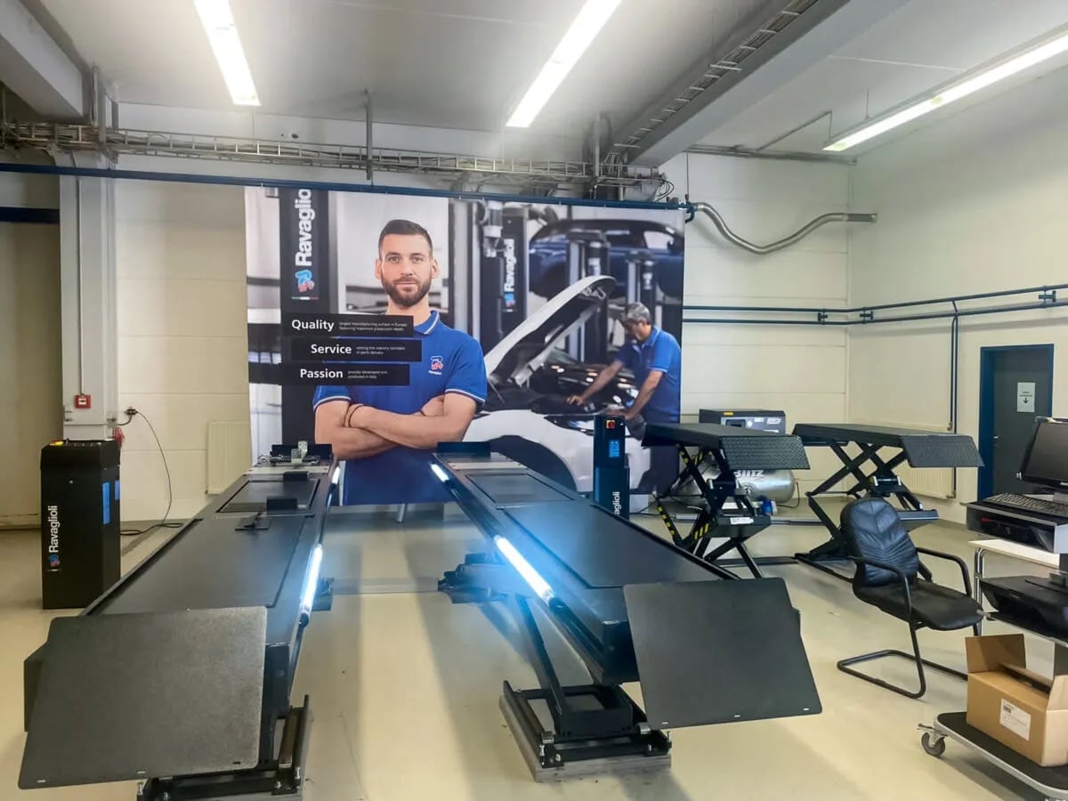 Werkstattinnenraum mit zwei flachen Fahrzeughebebühnen, großem Werbeplakat mit Mechaniker in blauer Arbeitskleidung, offen stehender Motorhaube im Hintergrund, Stuhl, Computerarbeitsplatz und Kartons auf dem Boden