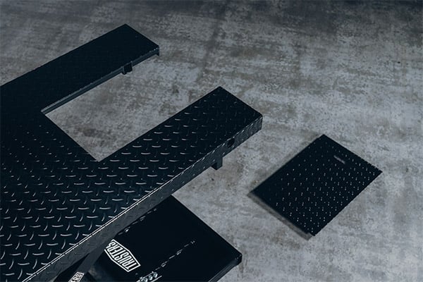 Black diamond-plate metal platform and ramp sections arranged on a concrete floor, with one smaller detached rectangular plate and a partially visible black toolbox underneath