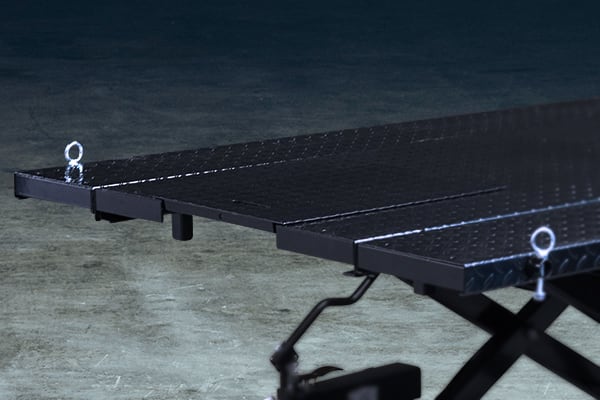 Close-up of a black metal scissor-lift platform with diamond-plate surface, two silver D-ring tie-downs at the edges, visible scissor mechanism beneath, on a concrete floor with a dark background