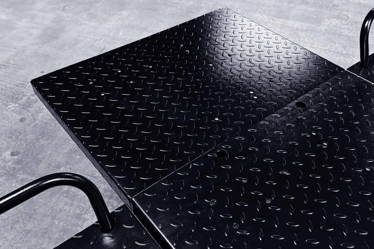 Close-up of black diamond-plate metal platform with raised tread pattern, two hinged panels joined by visible fasteners and curved black tubular handles, resting on a concrete floor