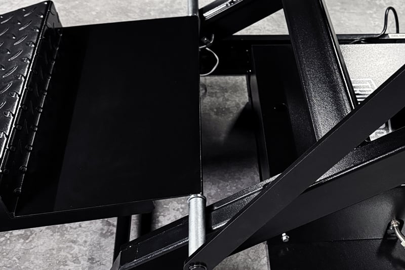 Detalle de piezas metálicas negras de un elevador tipo tijera: plataforma rectangular con chapa antideslizante, brazos articulados, pasadores cilíndricos y estructura sobre un suelo gris