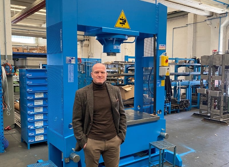 Mann in braunem Rollkragenpullover und graubraunem Sakko steht vor einer großen blauen Industriepresse in einer Werkhalle mit Regalen, Werkzeugkästen und Metallteilen im Hintergrund