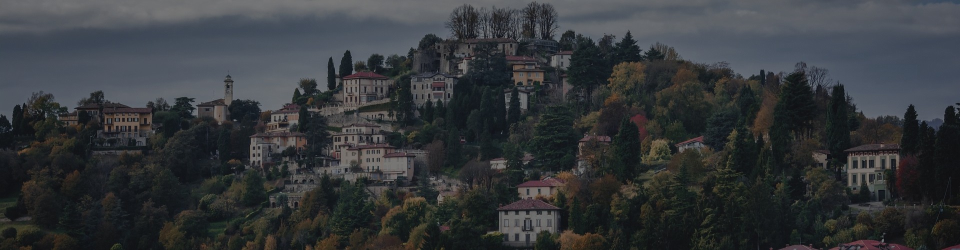 Panoramablick auf ein bewaldetes Hügeldorf mit historischen Häusern und Villen, Zypressen, herbstlich gefärbten Bäumen und einem Glockenturm unter einem dunklen, wolkigen Himmel