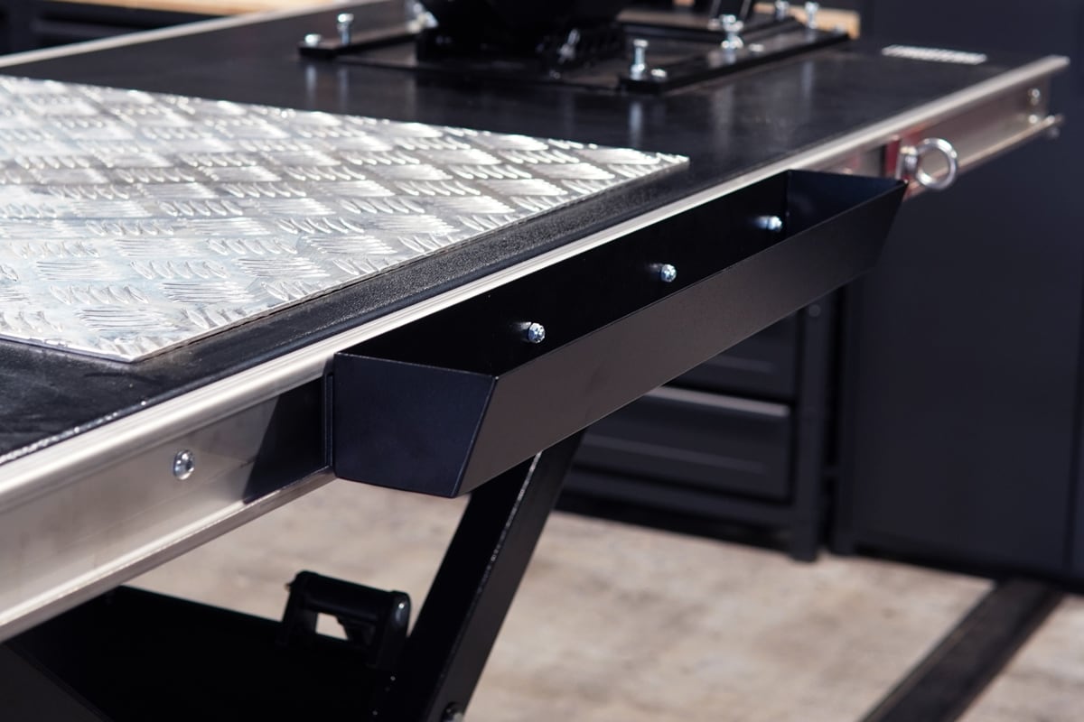 Nahaufnahme einer Werkbank mit Riffelblech-Auflage, Aluminiumkante und seitlich montiertem schwarzem Metallablagefach mit sichtbaren Schrauben, unscharfer Werkstatthintergrund.