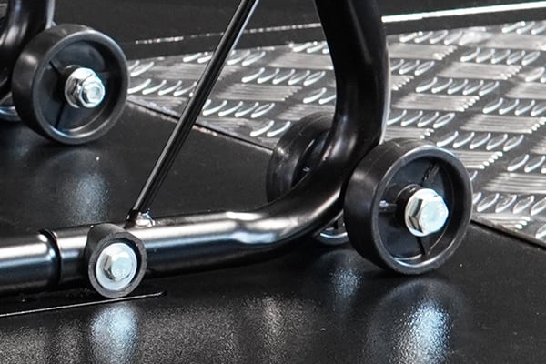 Detalle de un soporte metálico negro con ruedas y tornillos, apoyado sobre una placa de metal con patrón antideslizante y suelo reflectante
