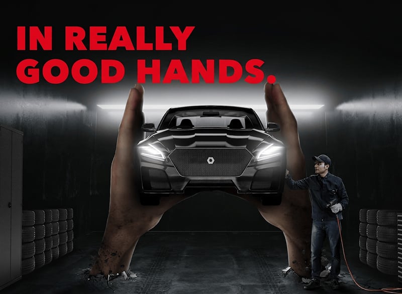 Dark automotive garage scene with giant hands cradling a black car front, mechanic in coveralls holding a tool at right, stacks of tires along walls and red headline text reading IN REALLY GOOD HANDS.