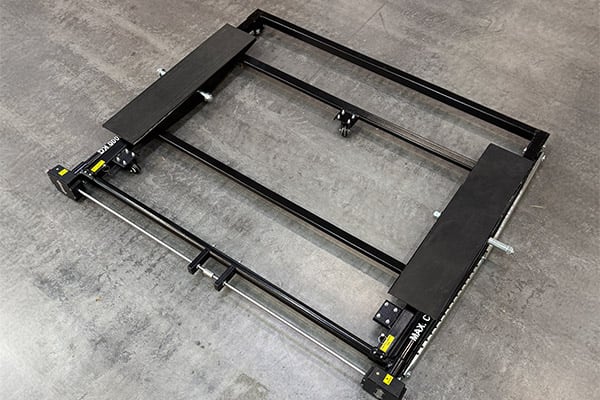 Schwarzer rechteckiger Metallrahmen mit zwei parallelen Stahlführungen, beweglichen Rollen, verstellbaren Spannschrauben und zwei gepolsterten Auflagen, liegend auf grauem Betonboden