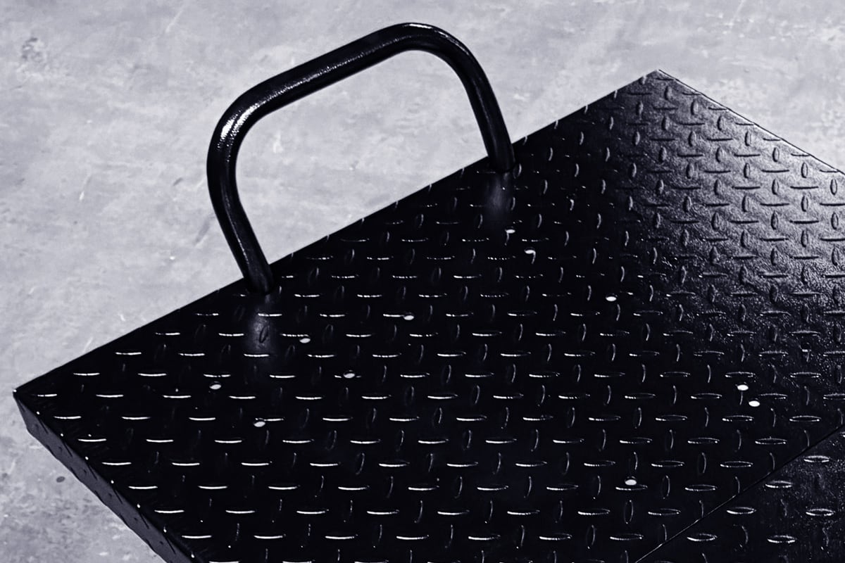Black diamond-plate metal platform with welded U-shaped handle viewed at an angle on a concrete floor