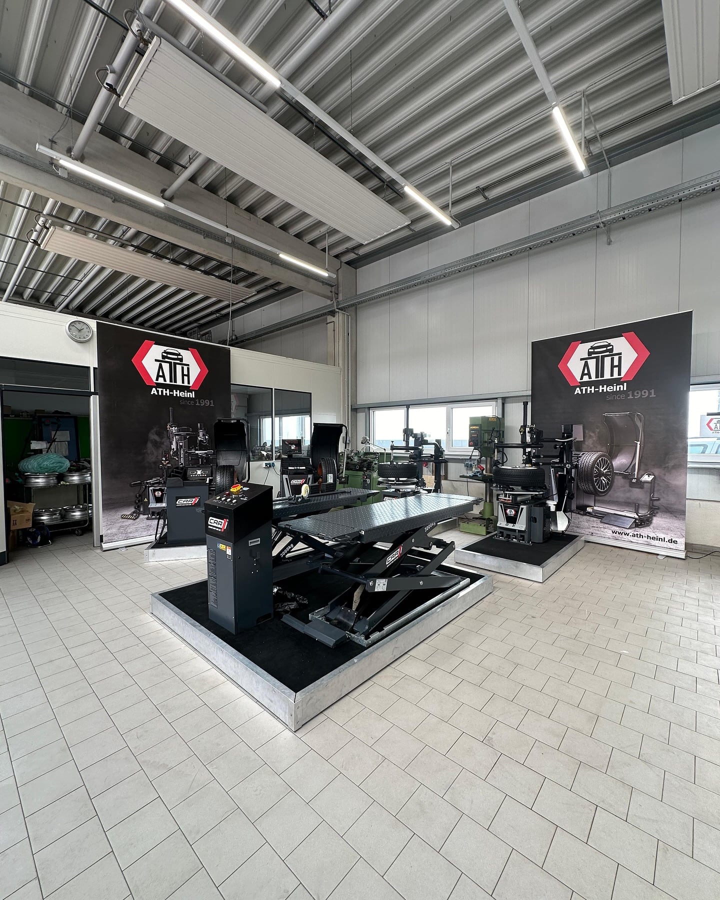 Innenraum einer Autowerkstatt mit zwei Scherenhebebühnen, Reifenmontiermaschinen, großen ATH-Heinl-Bannern an der Wand, hellem Fliesenboden und hohem Metallträgerdach mit hängenden Deckenleuchten