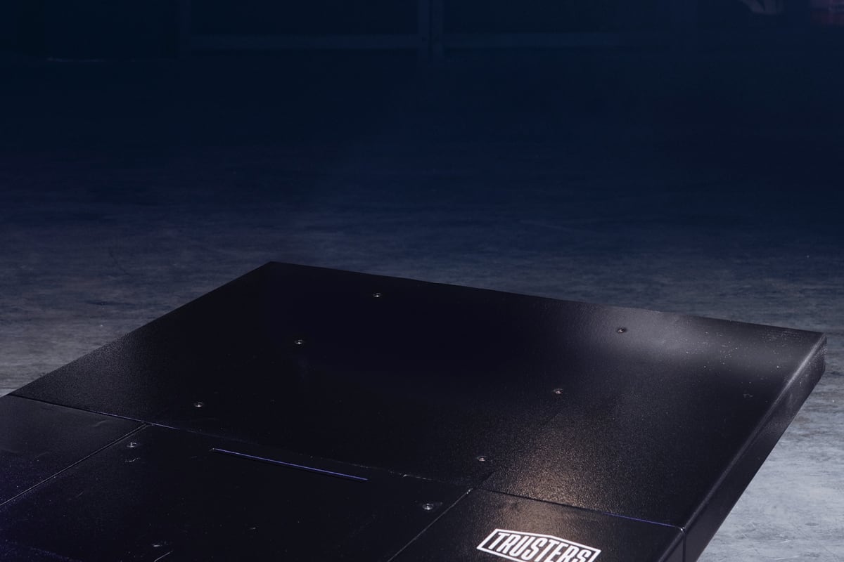 Low-angle view of a black metal platform with visible screws and a white TRUSTERS label on the corner, resting on a dimly lit concrete floor