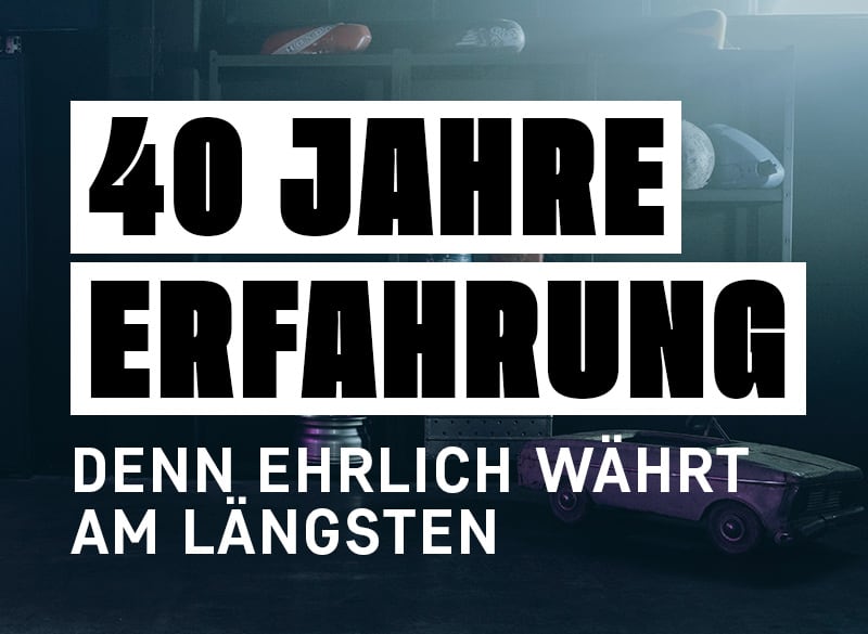 Weiße fette Schrift 40 Jahre Erfahrung und darunter Denn ehrlich währt am längsten vor dunklem Werkstatt-/Garagenhintergrund mit altem Auto, Regalen und Werkzeug