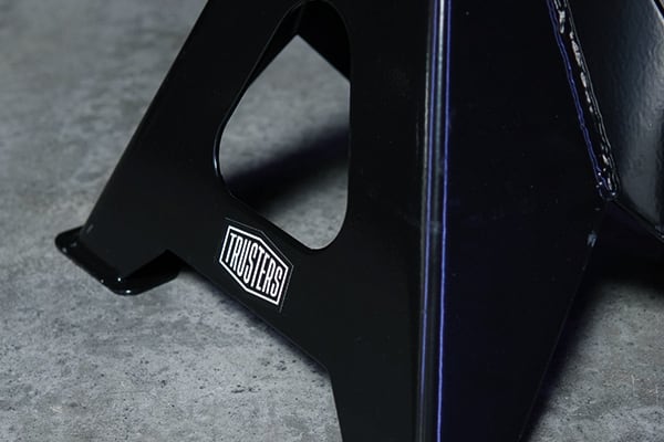 Nahaufnahme eines schwarzen Metall-Unterstellbocks mit ausgeschnittenem Tragegriff, weißer Aufkleber mit Schriftzug TRUSTERS, auf grauem Betonboden