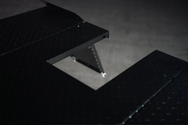 Black metal diamond-plate platform with a rectangular cutout showing a folding support leg and white foot against a blurred gray background