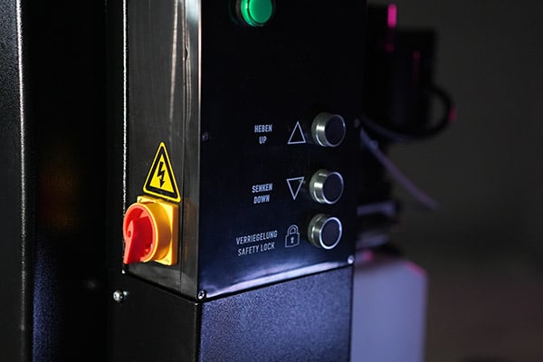 Close-up of a machine control panel showing a red emergency stop switch with a yellow high-voltage warning triangle, a green indicator light, and three metal buttons labeled HEBEN/UP, SENKEN/DOWN and VERRIEGELUNG/SAFETY LOCK