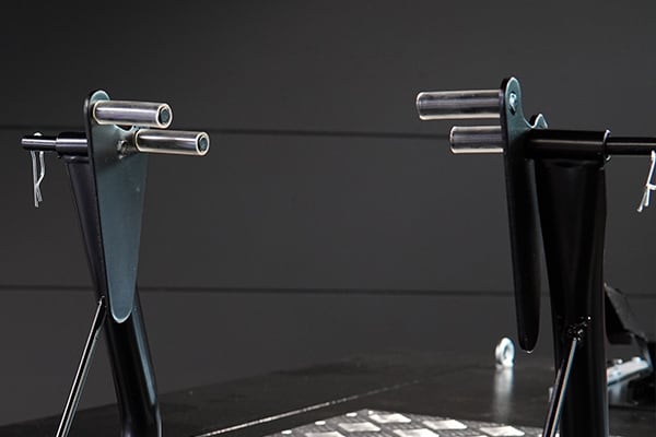Dos soportes metálicos negros enfrentados con brazos superiores que sostienen ejes cilíndricos plateados horizontales sobre una mesa de trabajo oscura y fondo gris uniforme
