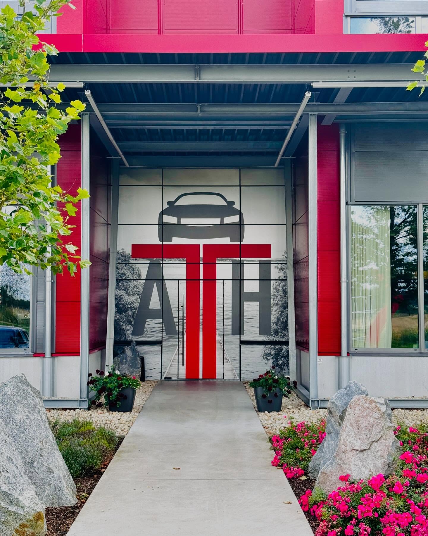Eingang zu einem Gebäude mit roter Fassade und Metallvordach, Glasfront mit stilisierter Autosilhouette über großen roten Buchstaben, breiter Betonweg, Kübelpflanzen, pinke Blumenbeete und dekorative Zierfelsen