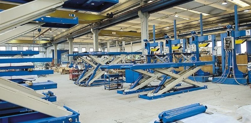 Große Industriehalle mit mehreren blauen hydraulischen Scherenhebebühnen und Arbeitsplattformen, hohem Hallendach mit Laufkran, Werkzeugen und Lagerflächen auf Betonboden, helle Fensterreihe im Hintergrund